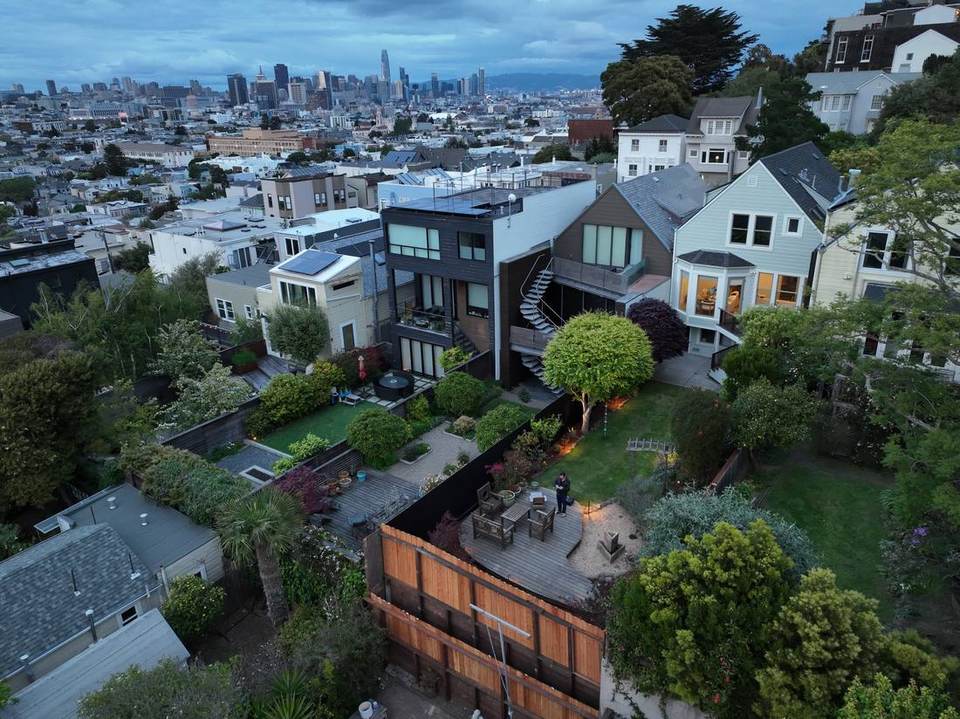 Aerial view at dusk of deep backyards behind a row of San Francisco houses, with downtown visible in the distance